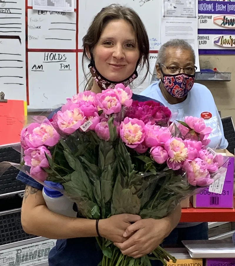 Abby holding a big armful of pink peonies in the back room at Trader Joe’s in 2020, both employees masked.
